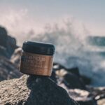 Close-up of CBD infused gel jar on rocks by the sea, showcasing product against a scenic ocean backdrop with splashing waves.