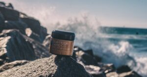 Close-up of CBD infused gel jar on rocks by the sea, showcasing product against a scenic ocean backdrop with splashing waves.
