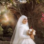 A beautiful bride in a white dress and veil holds a bouquet, smiling in a rustic outdoor setting.